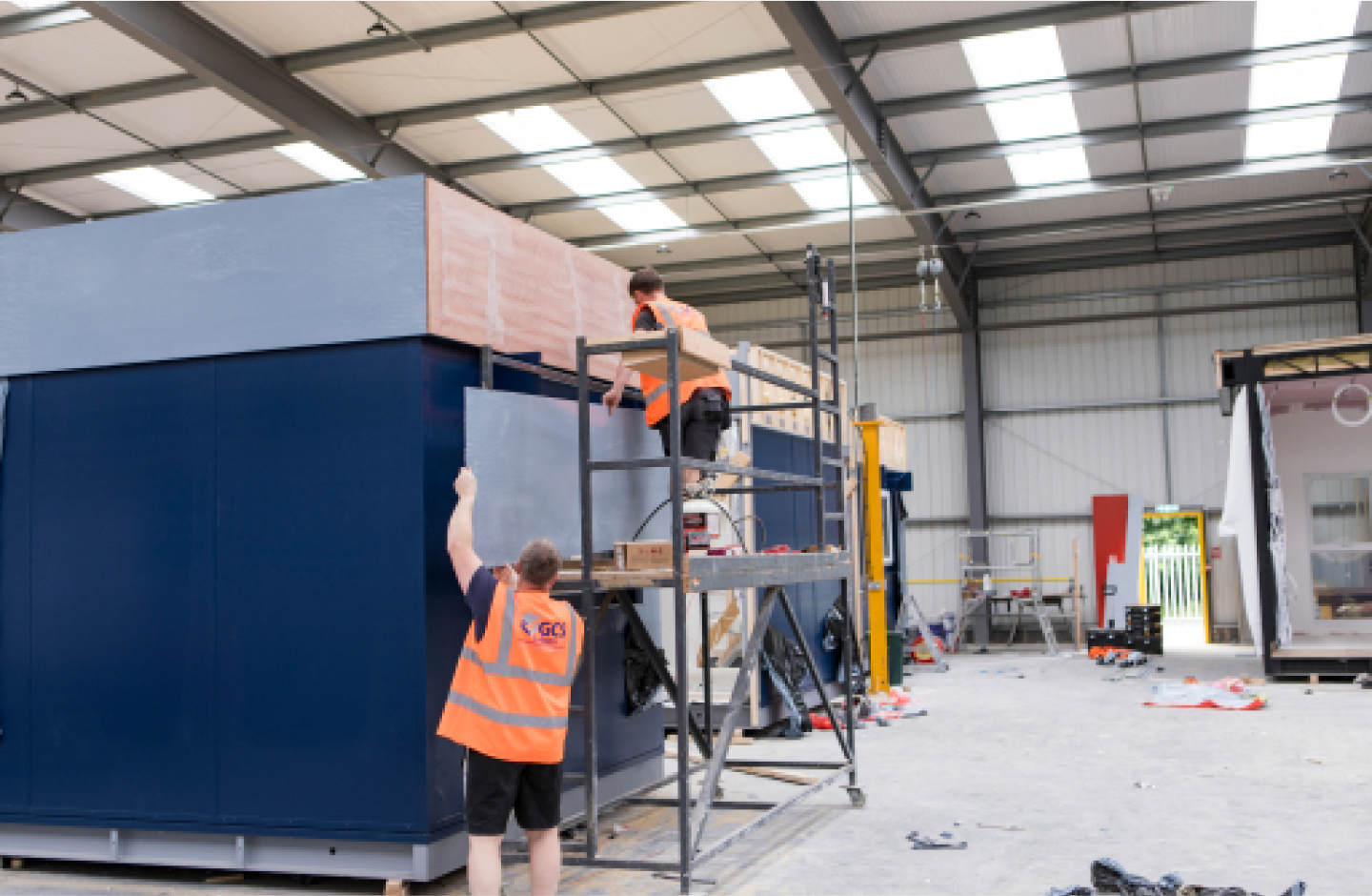 two people work on the side of blue modular building in a factory